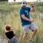 A dad dancing in a field with his young daughter. He's wearing a hat and teal shirt, while she looks up at him. A fun moment of house painting break!