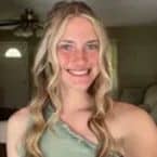 Smiling woman with blonde wavy hair, wearing a green top. A glimpse of a cozy home interior can be seen in the background. Consider house painting to refresh your space.