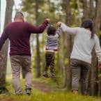 A family of three enjoys a walk in the woods; parents hold their child up between them, swinging them along the path. Trees surround them in a natural, earthy setting.