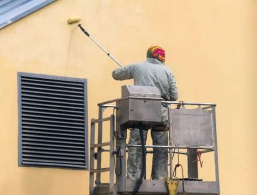 A house painting professional on a lift uses a roller to apply fresh paint to the exterior wall of a building. The building is light yellow, and a window with black slats is visible.