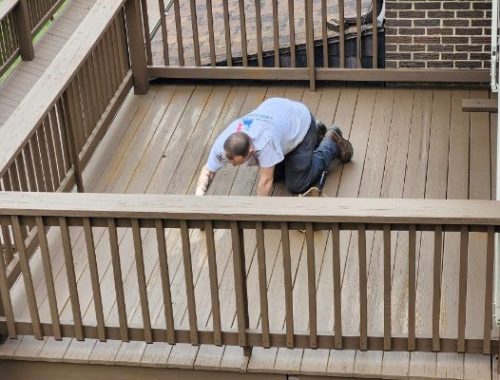 A man on his knees stains a wooden deck brown. He is working on house painting, visible from the railing and ramp in the background.