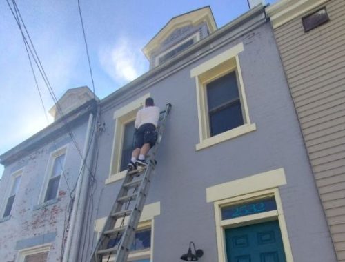 A man on a ladder paints the exterior of a two-story gray house with yellow trim. The house has a blue door with the number 2532 above it. Power lines are visible against a blue sky.