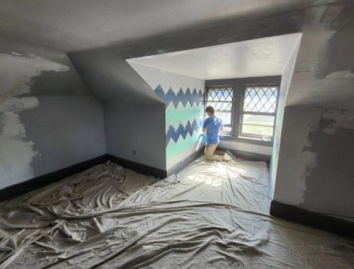 Painter renovates an attic room, applying a teal geometric design to the wall. The room is prepped with drop cloths and features a window with diamond-shaped panes.